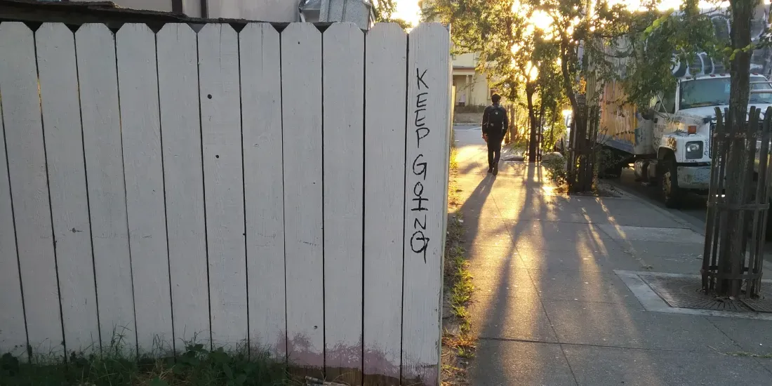 Old white picket fence with the words "keep going" written in marker on the edge. A person walks away along the sidewalk, toward a setting sun.