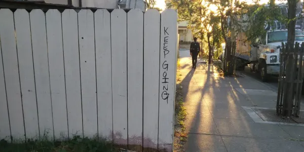 Old white picket fence with the words "keep going" written in marker on the edge. A person walks away along the sidewalk, toward a setting sun.